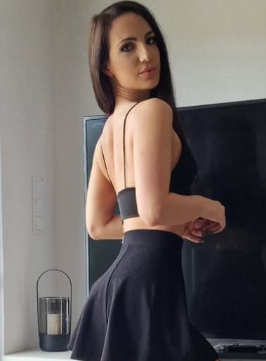 Kristy Black se fotí v černém crop topu a černé krátké sukni, stojící před televizí.