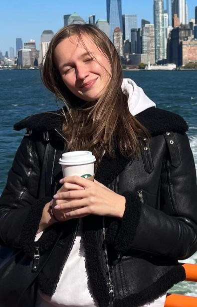 Stacy Cruz takes a photo in front of her city and dressed in white hoodie and black leather jacket while holding a coffee cup in her hand.