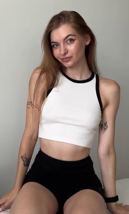 Kortie se fotí, jak sedí na posteli a oblečená v sexy bílém crop topu a černých kraťasech.