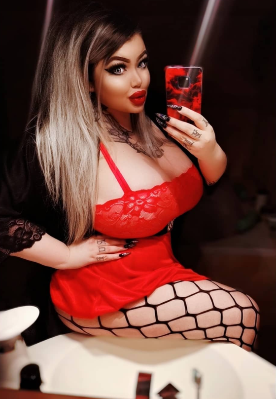 Rebecca Randallová se fotí v koupelně a je oblečená v sexy červených šatech a černé bundě.